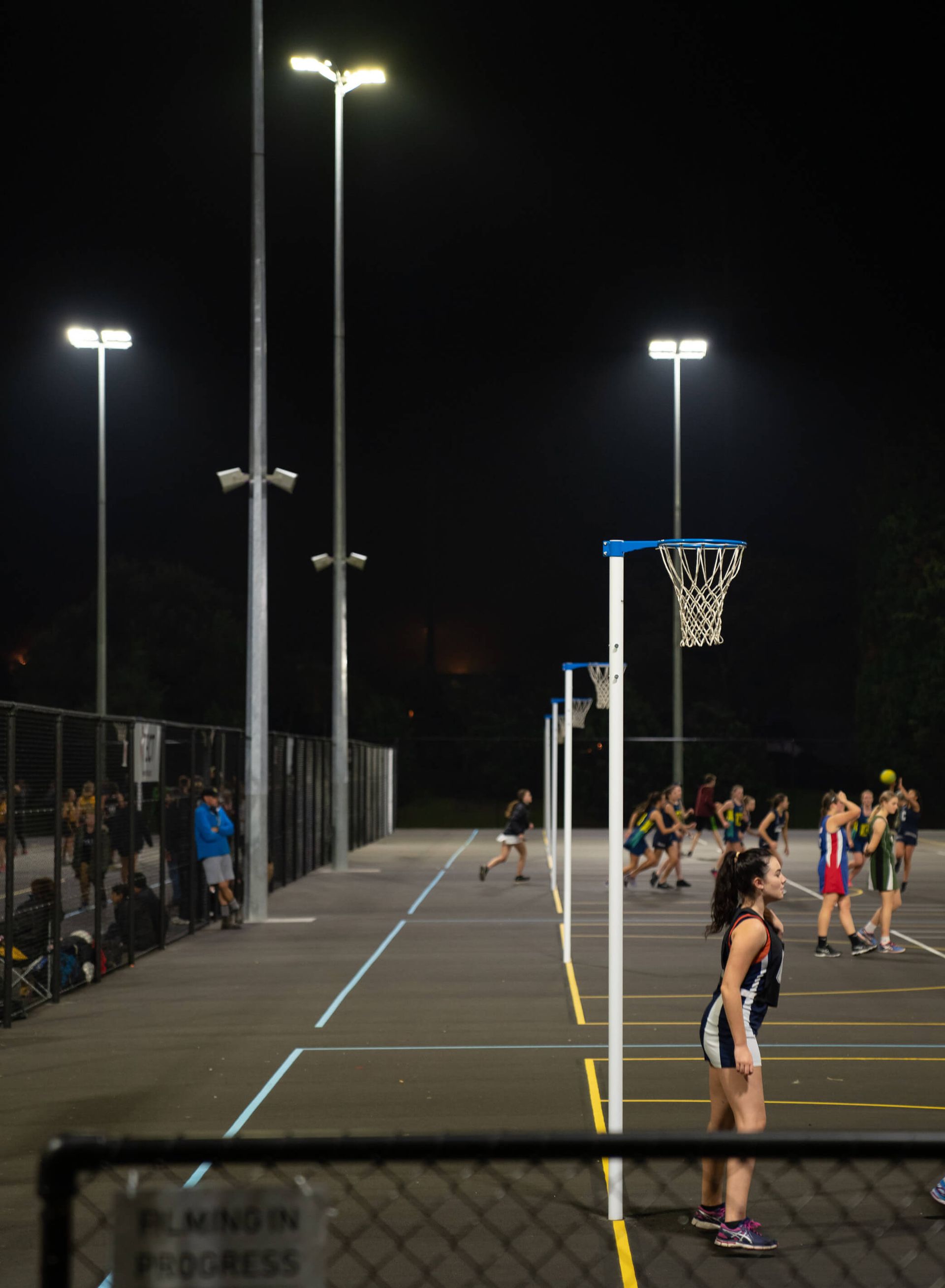 Netball game at night under bright lights. Players in uniforms on a court with hoops; spectators along the side.