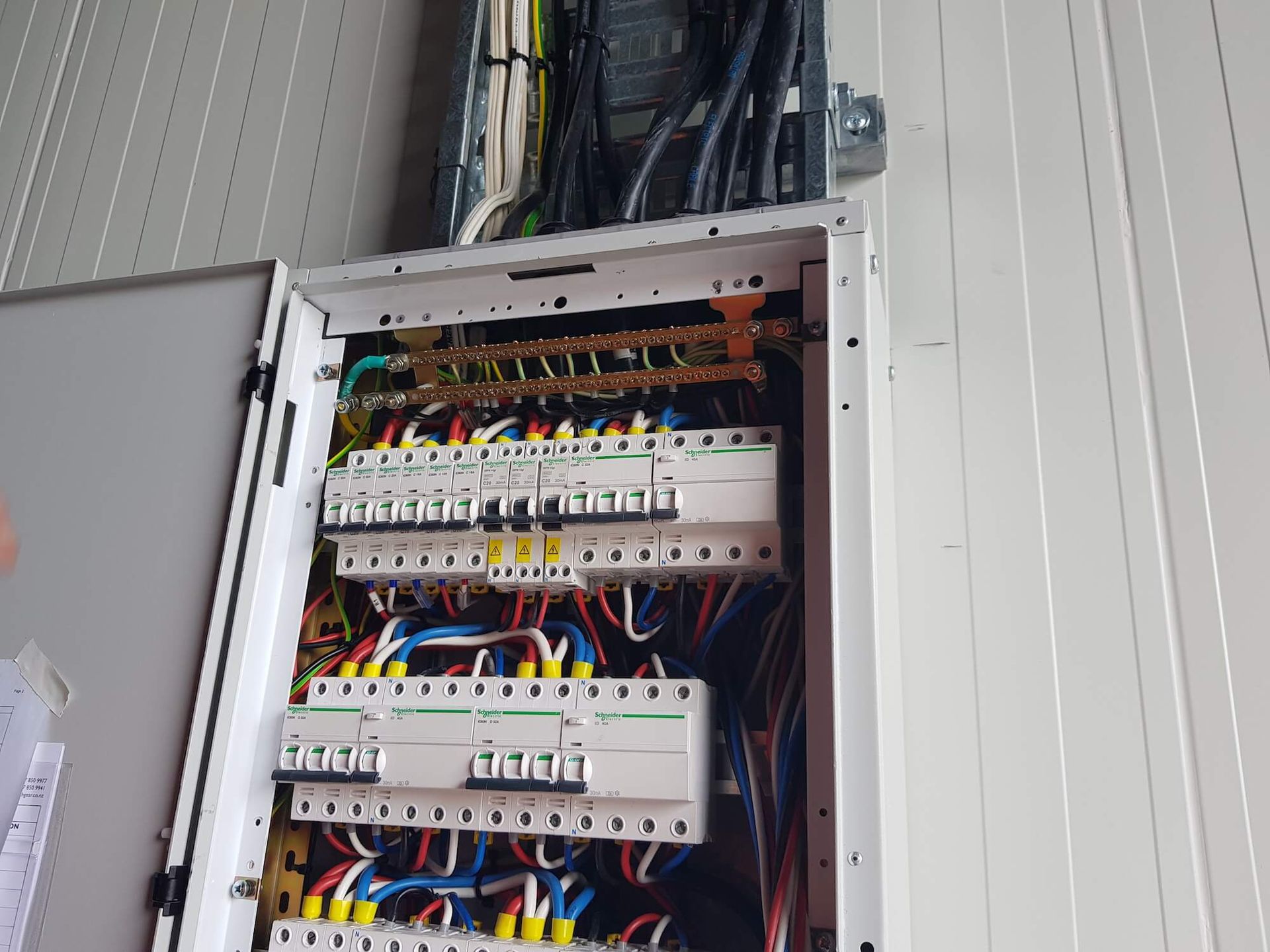 Electrical panel with circuit breakers and wiring, mounted on a white wall. Wires are color-coded.