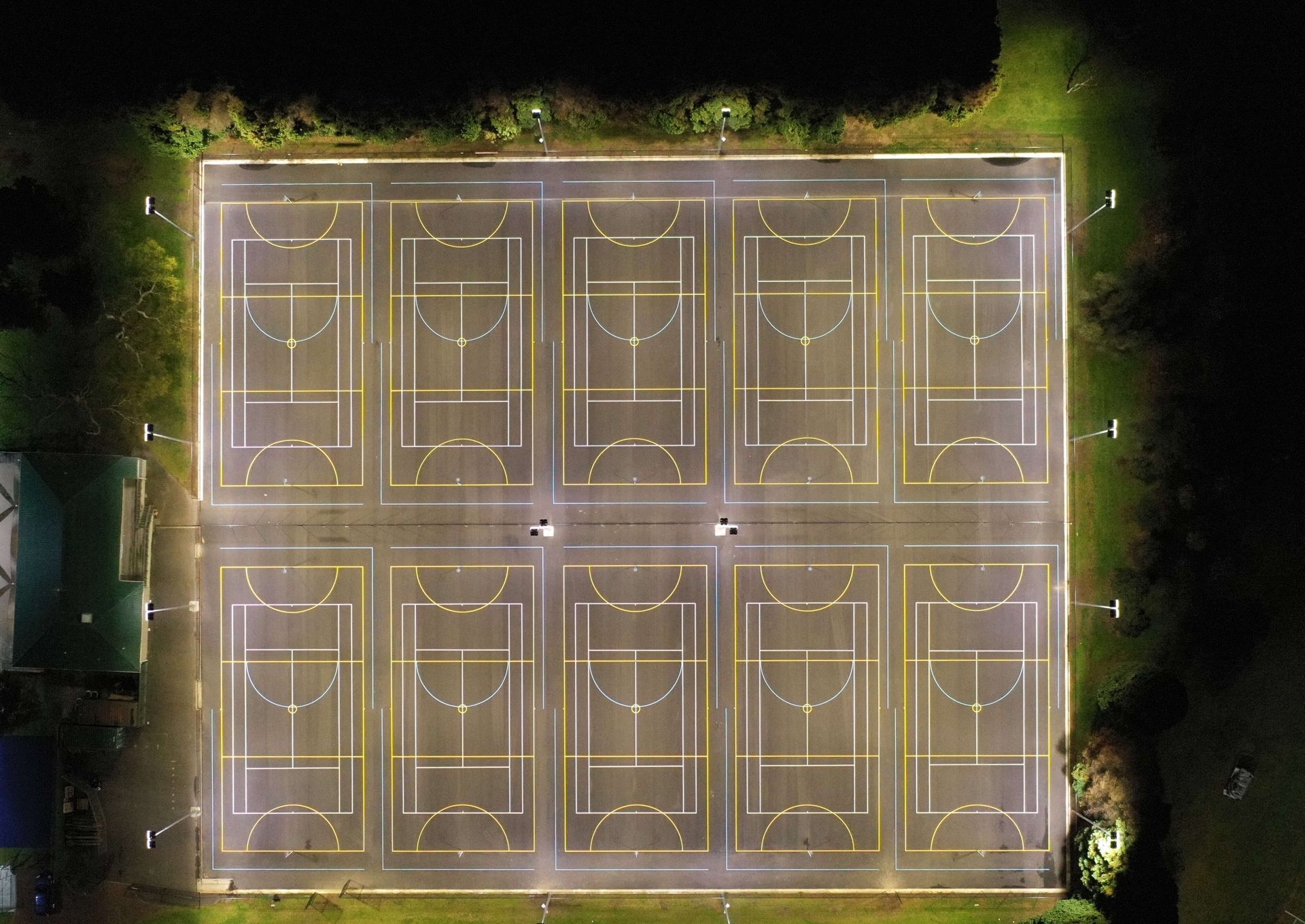 Overhead view of a rectangular outdoor court illuminated at night, with lines for multiple sports.