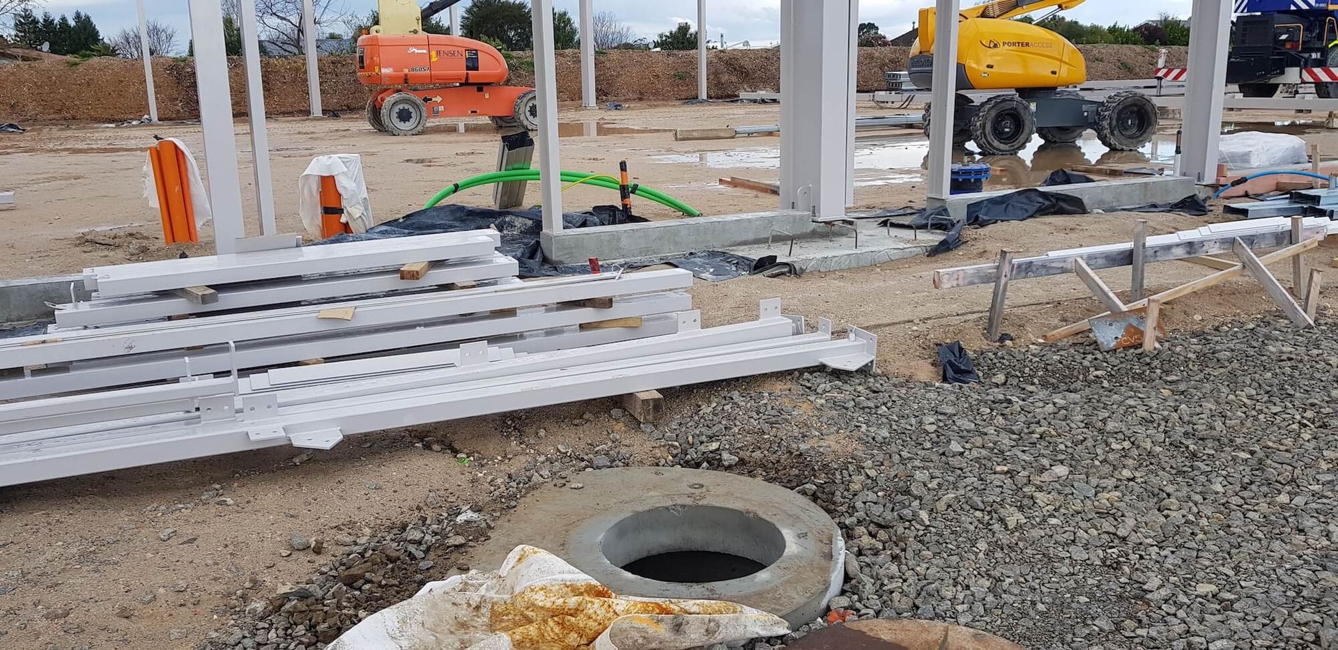 Construction site with white concrete pillars and various equipment including an orange lift and a yellow excavator, on a gravel ground.
