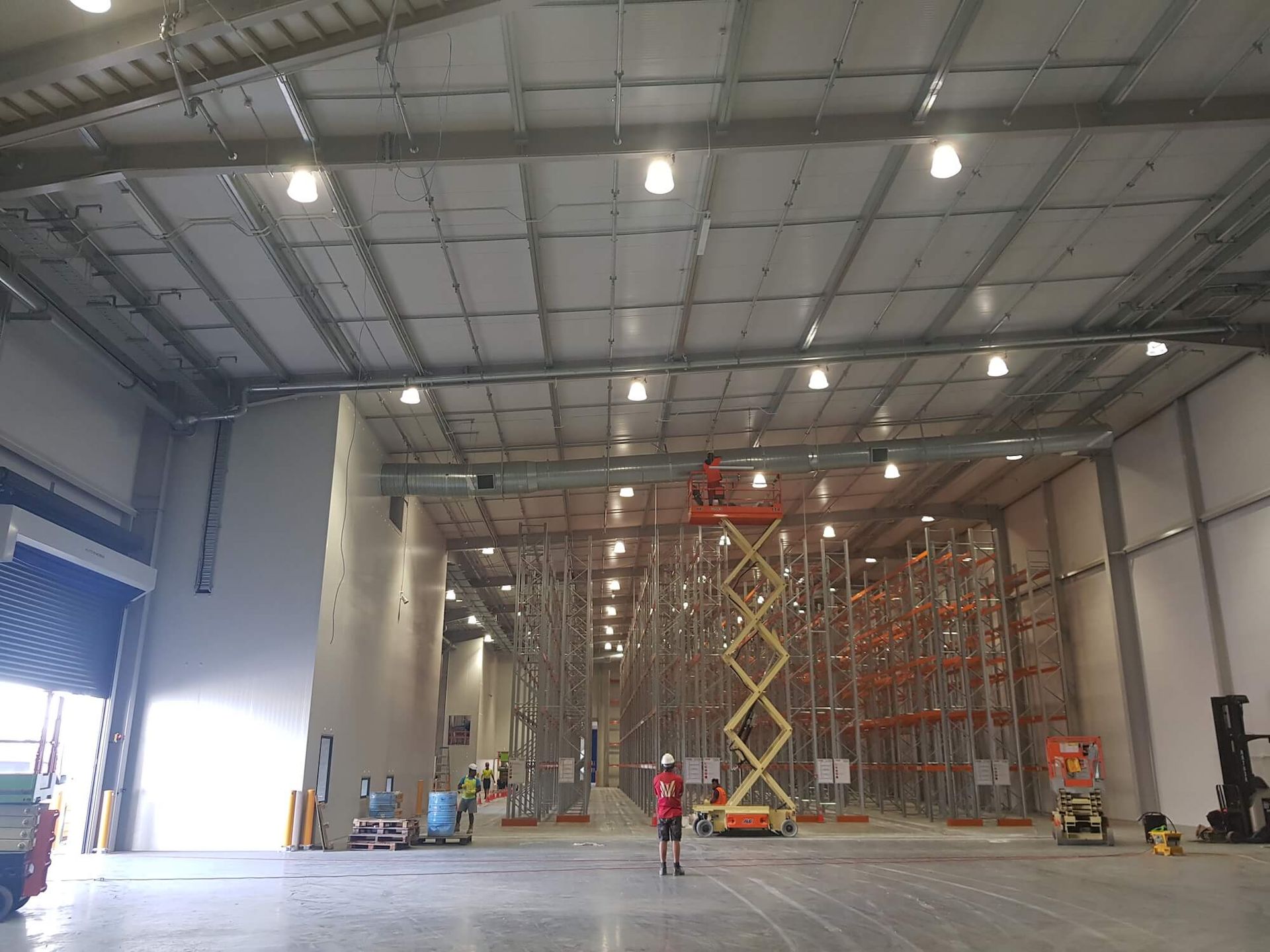 Inside a warehouse: worker on a scissor lift, installing fixtures. Racks line the background.