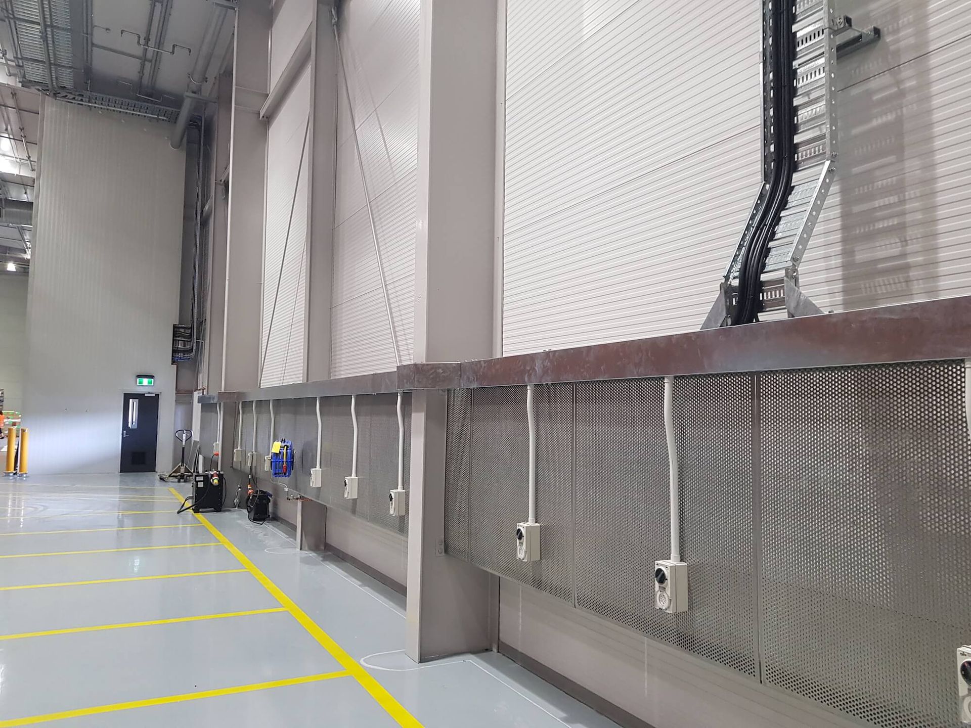 Long industrial loading dock with metal barriers, electrical outlets, and overhead cable management. Yellow painted floor lines lead into the space.
