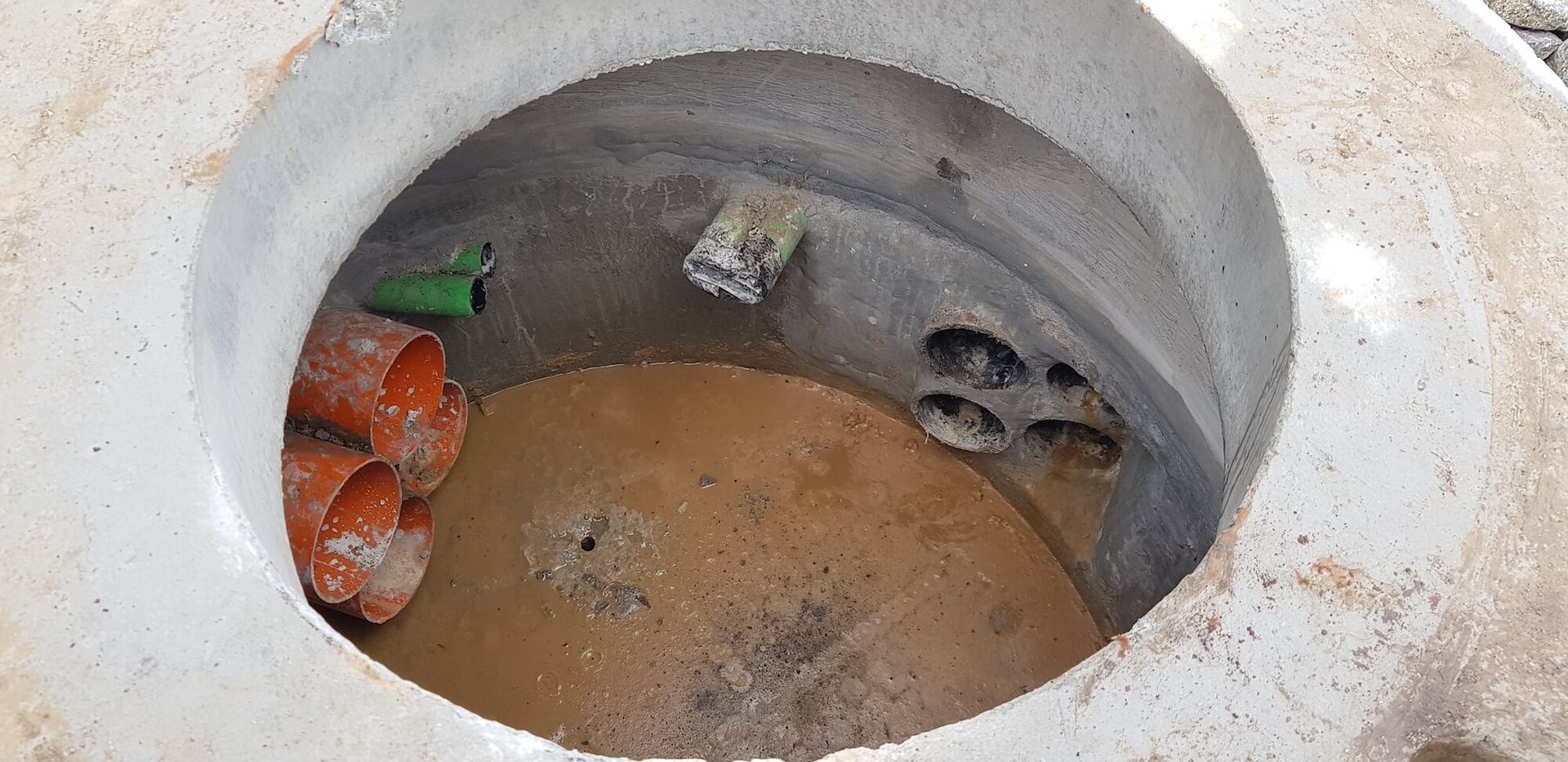 Looking down into a concrete manhole containing orange and green pipes. There are a few dark holes in the side of the manhole.