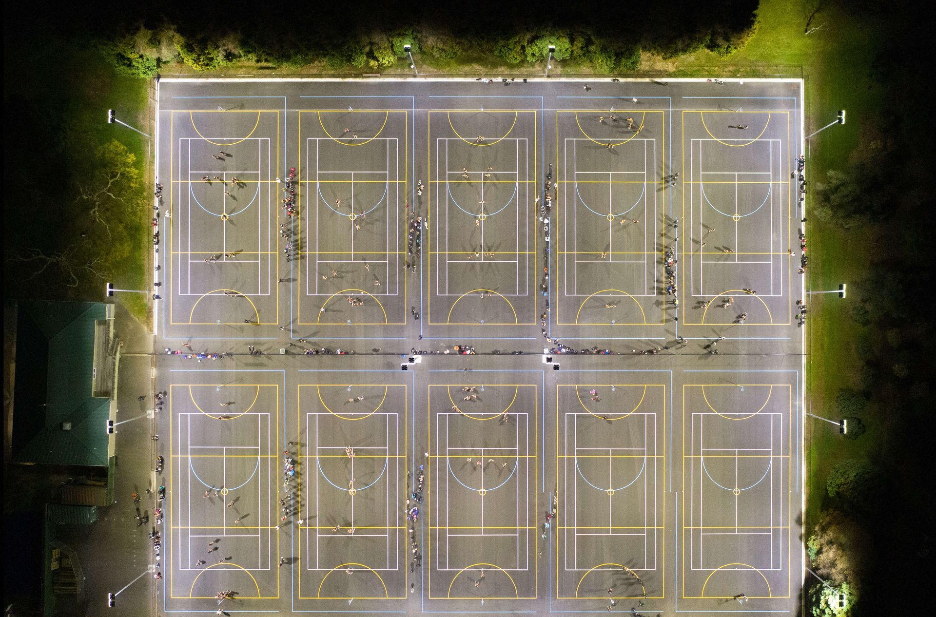 Overhead night view of a lit outdoor sports court with basketball and other game markings, people playing.