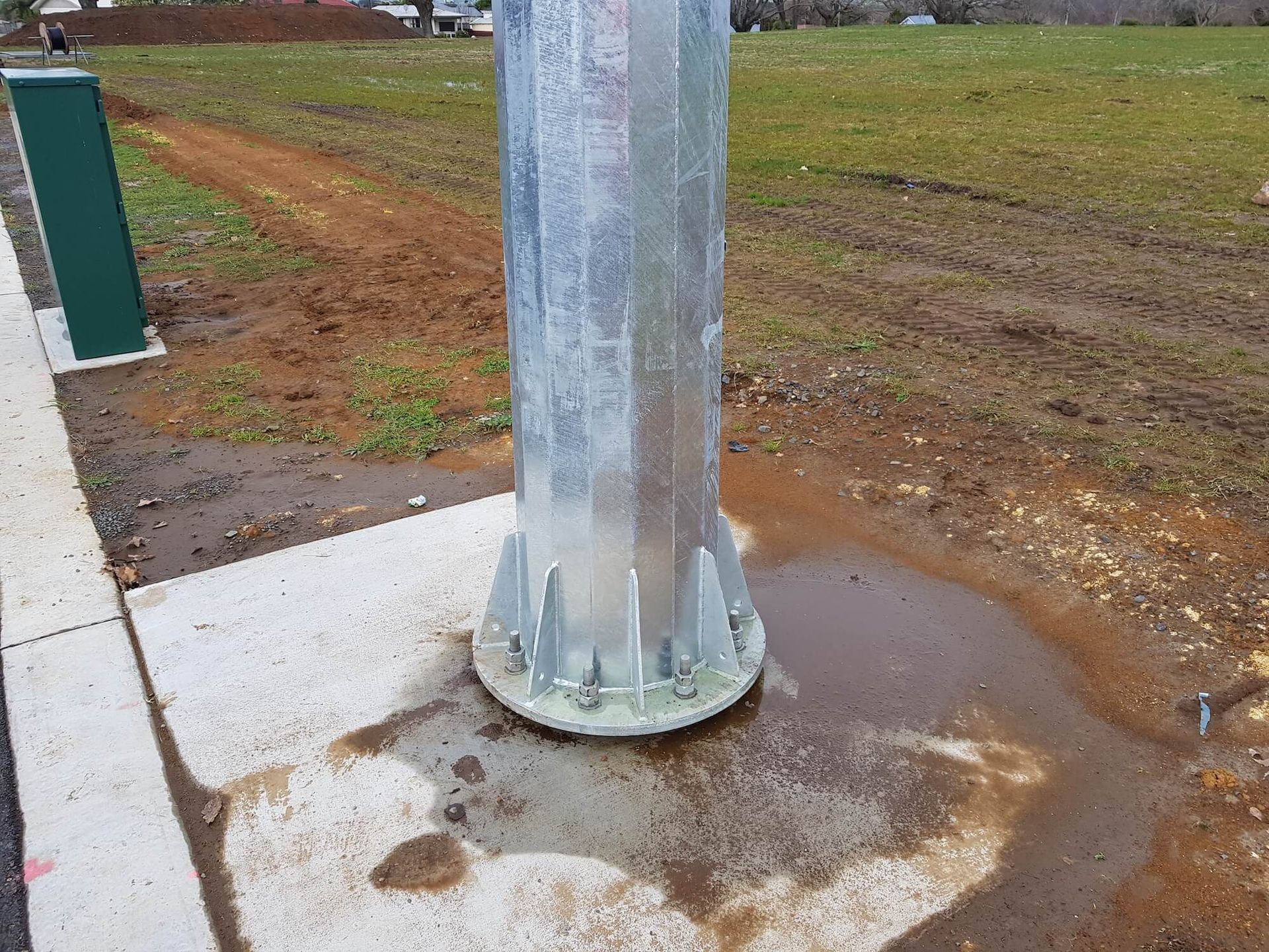 A metallic light post secured to a concrete base, surrounded by wet soil and a green utility box.
