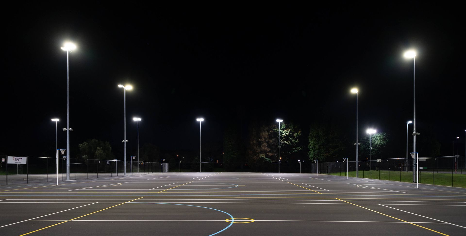 An empty, dark parking lot at night, illuminated by bright streetlights. Pavement lines and reflectors are visible.