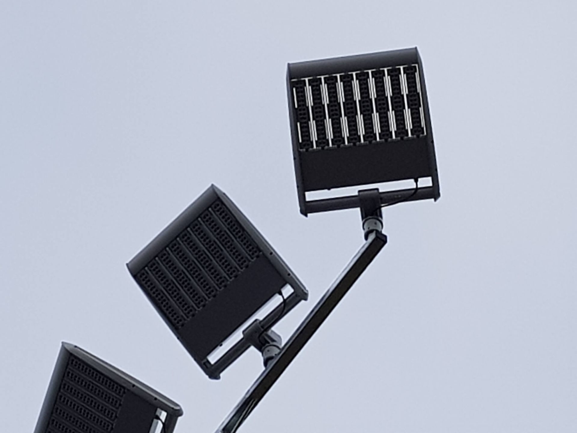 Three dark square LED floodlights mounted on a black pole against a light gray sky.
