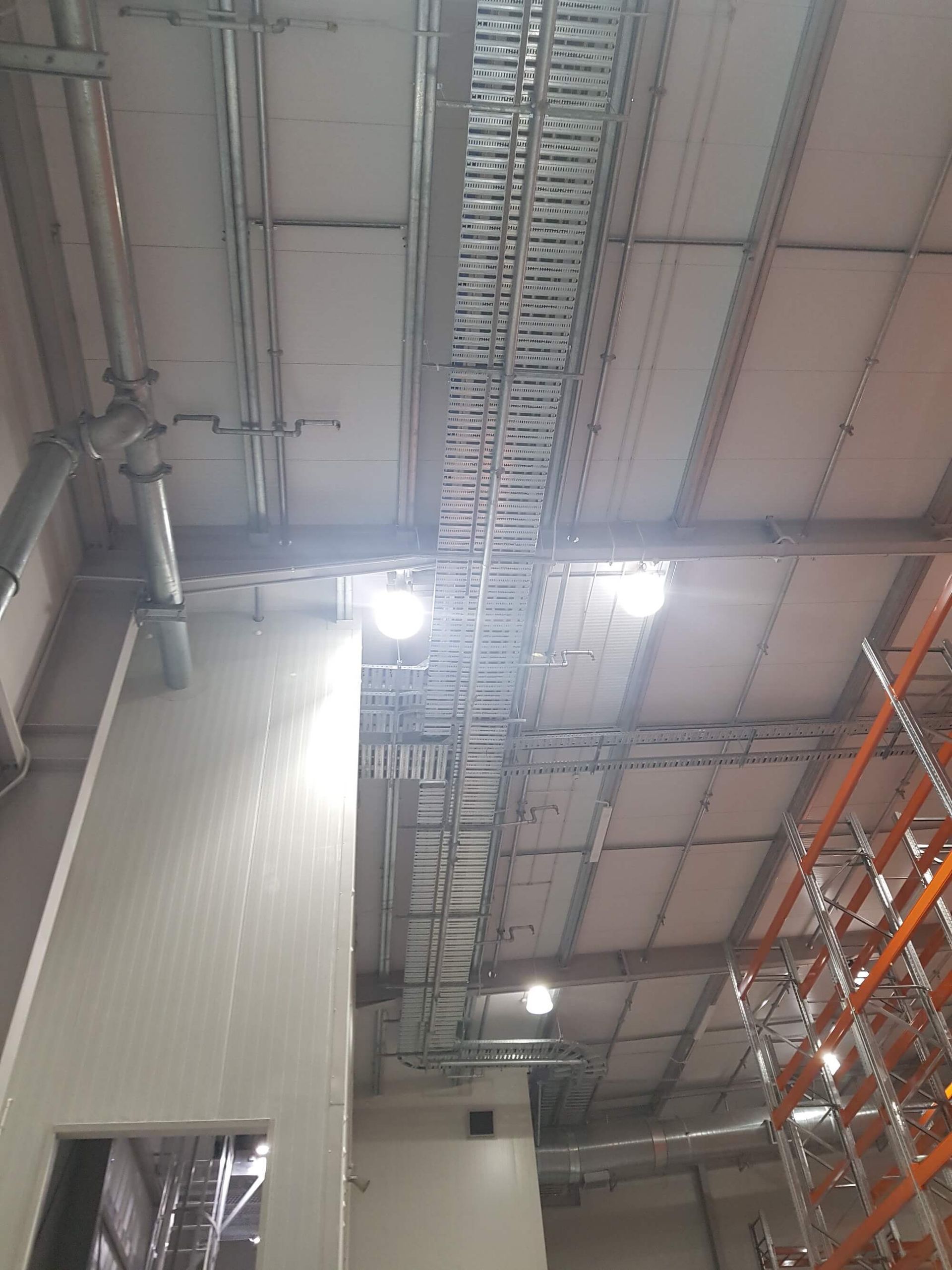 Warehouse ceiling with metal cable trays, lighting, and insulation panels. Metal support beams and storage racks are visible.