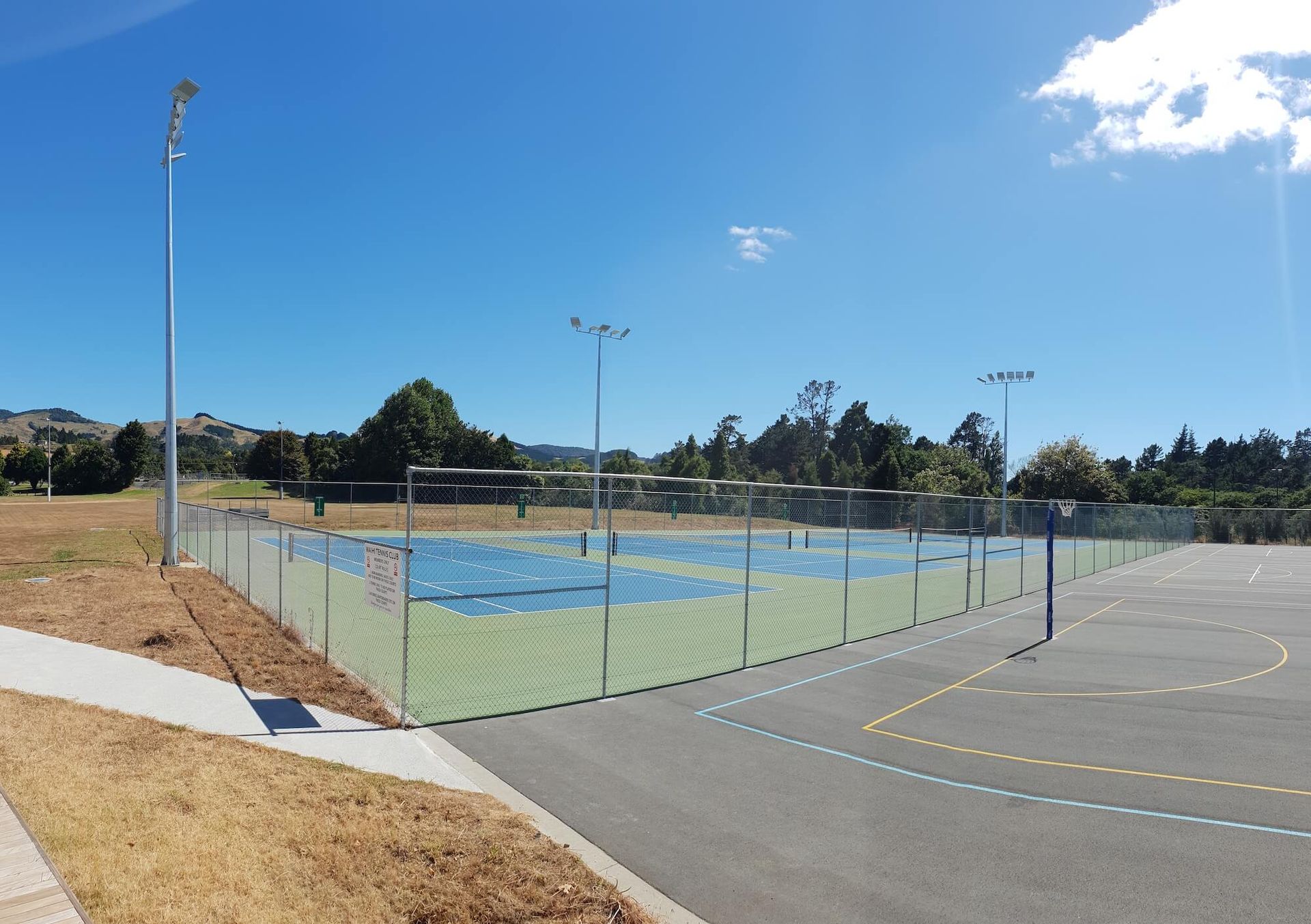 Outdoor sports court with blue and green surface, surrounded by a fence and pathway, under a blue sky. Outdoor sports court with blue and green surface, surrounded by a fence and pathway, under a blue sky.