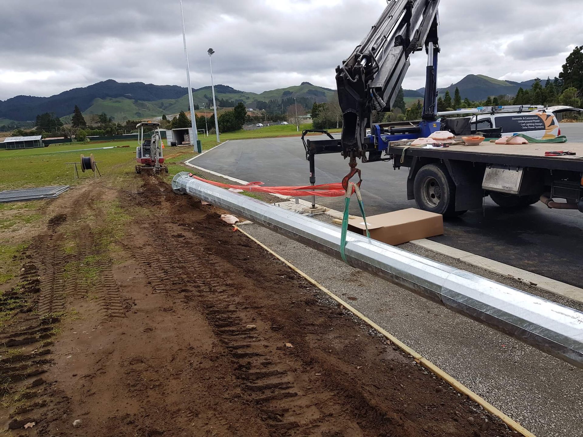 A truck crane lifting a long metal beam next to a curb on a roadside with hills in the background.