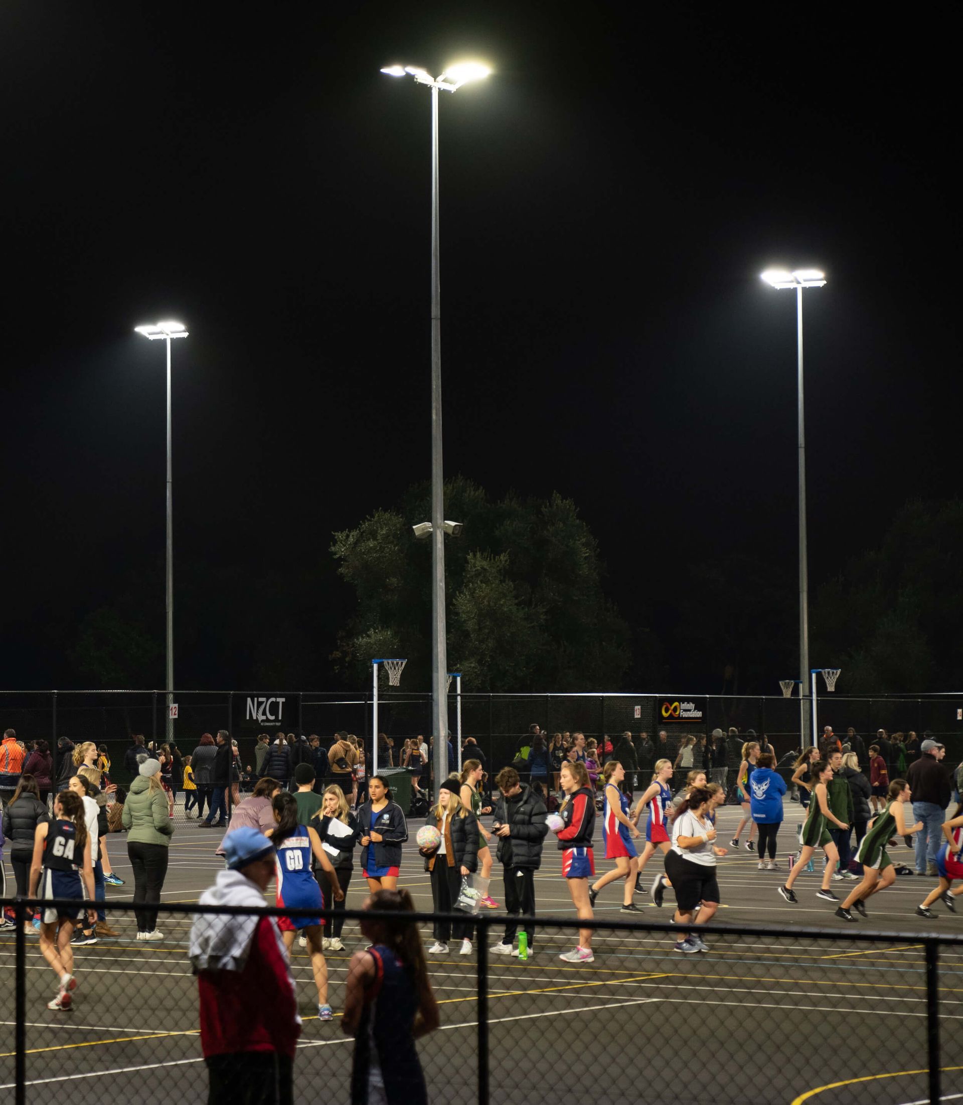 Netball game at night under bright floodlights, with numerous players on the court and spectators watching.