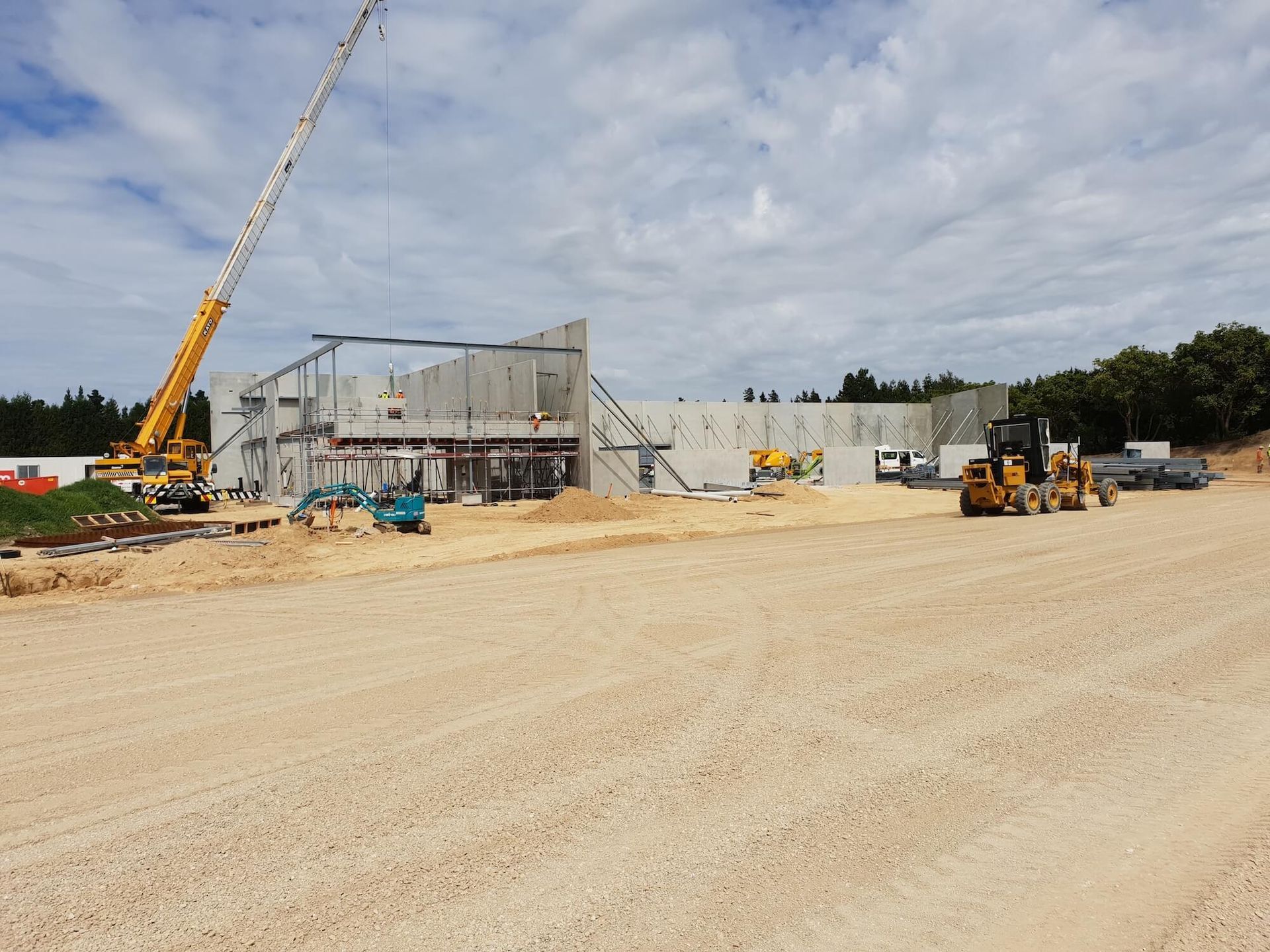 Construction site with crane and heavy machinery; unfinished concrete building under construction. Cloudy sky and dirt ground.