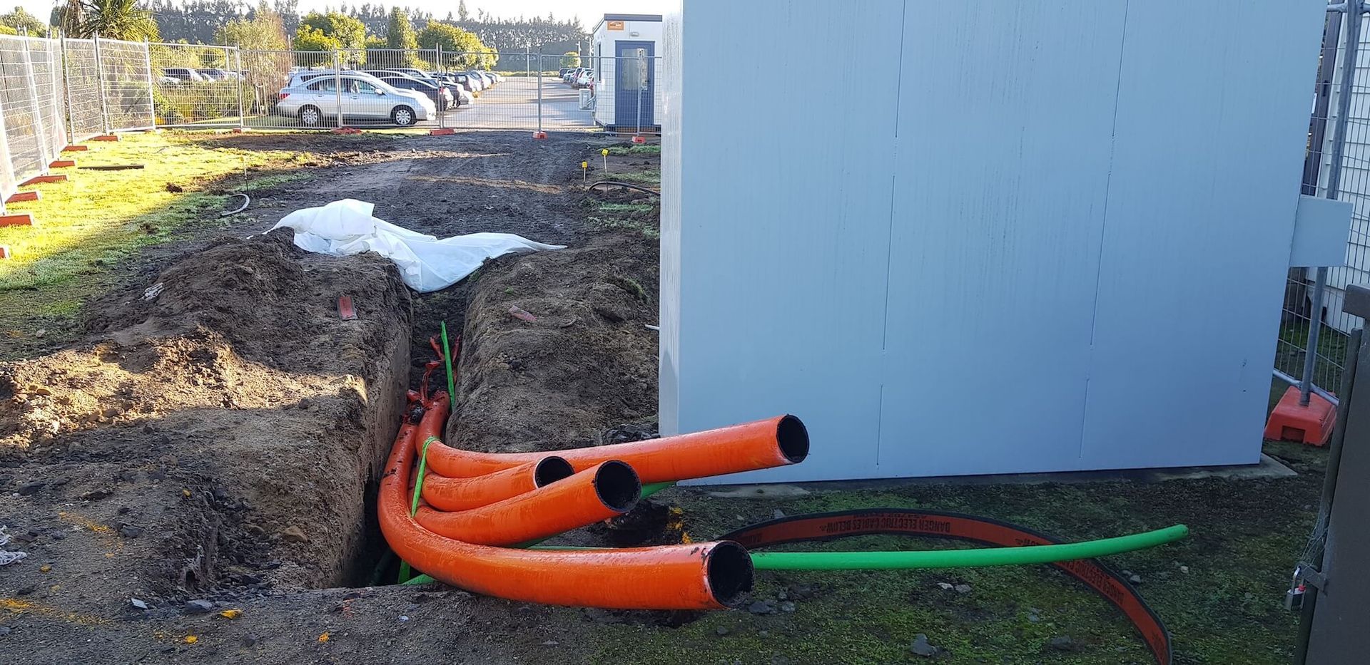 Construction site with orange and green conduit pipes emerging from a trench near a building. Cars are parked in the background.
