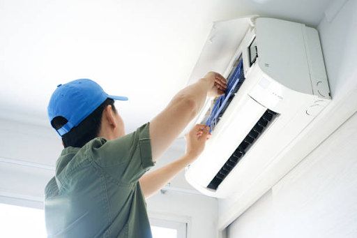 A person in a blue cap and green shirt is servicing a white air conditioner mounted on a wall, remov