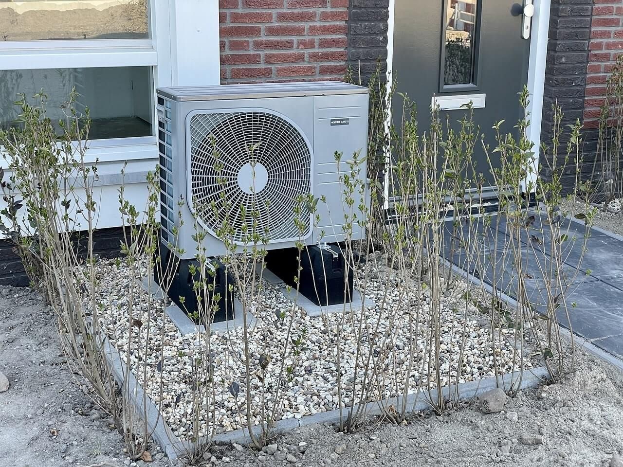Air source heat pump unit on a gravel bed, surrounded by young bare-branched shrubs, near a house en