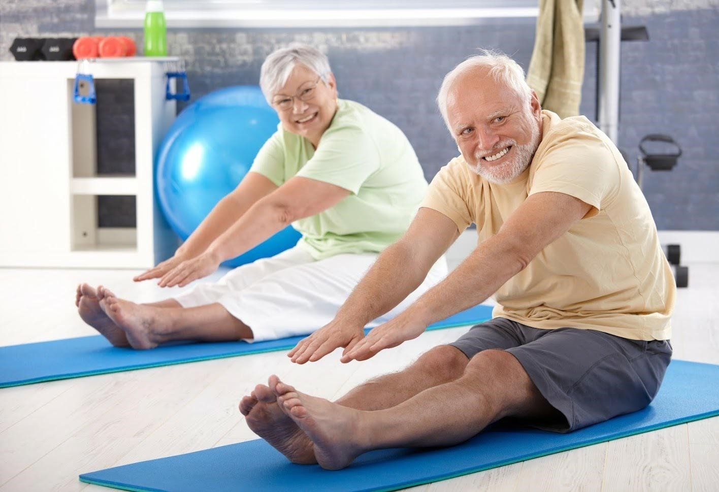 Stretching and Yoga – Philadelphia, PA – Advanced Physical Therapy Two people, seated on mats, stretching toward their toes. Light-colored clothing, happy expressions, indoor setting.