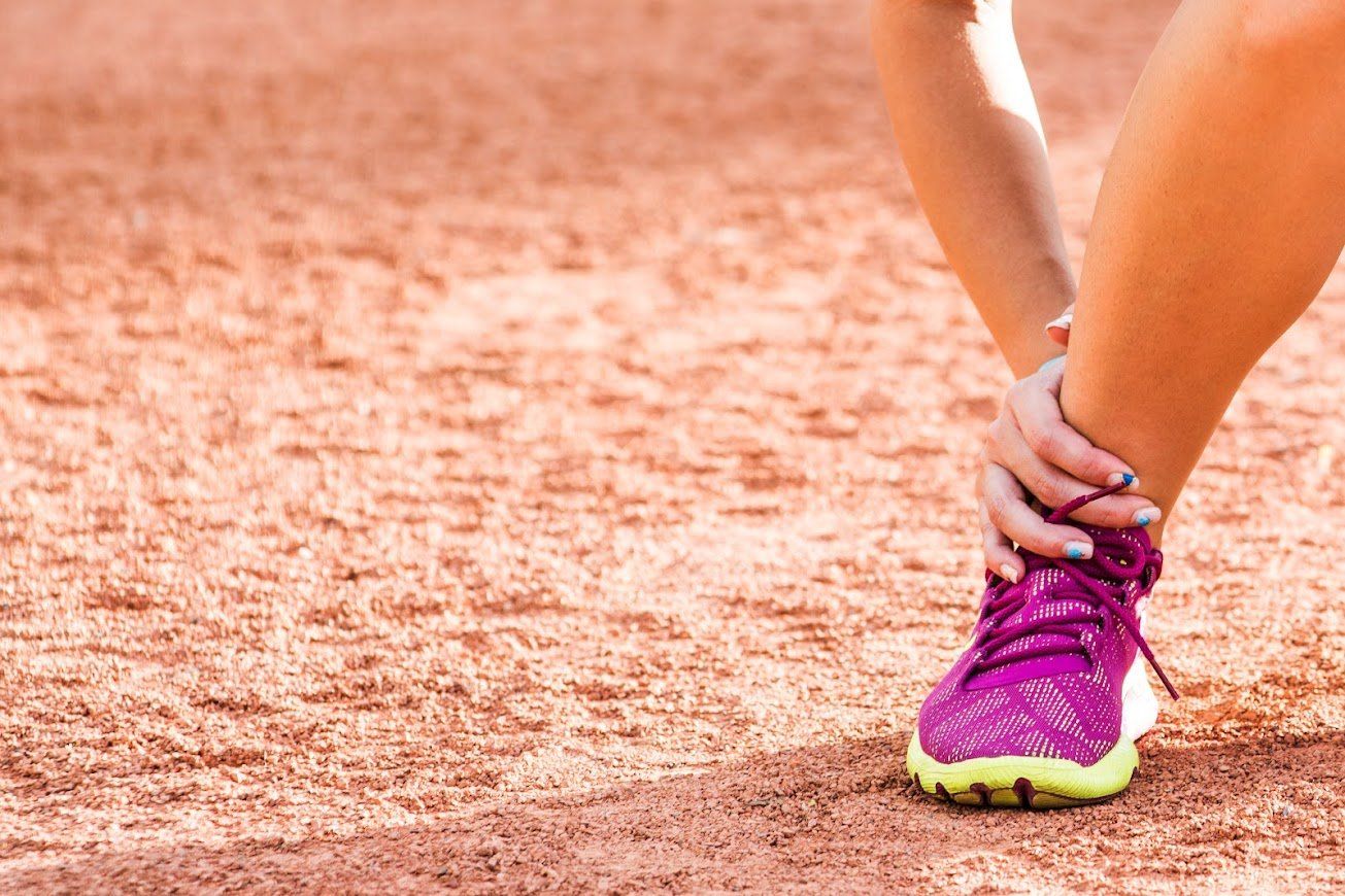 A person holding their ankle in pain while wearing bright pink sneakers on a clay sports court.