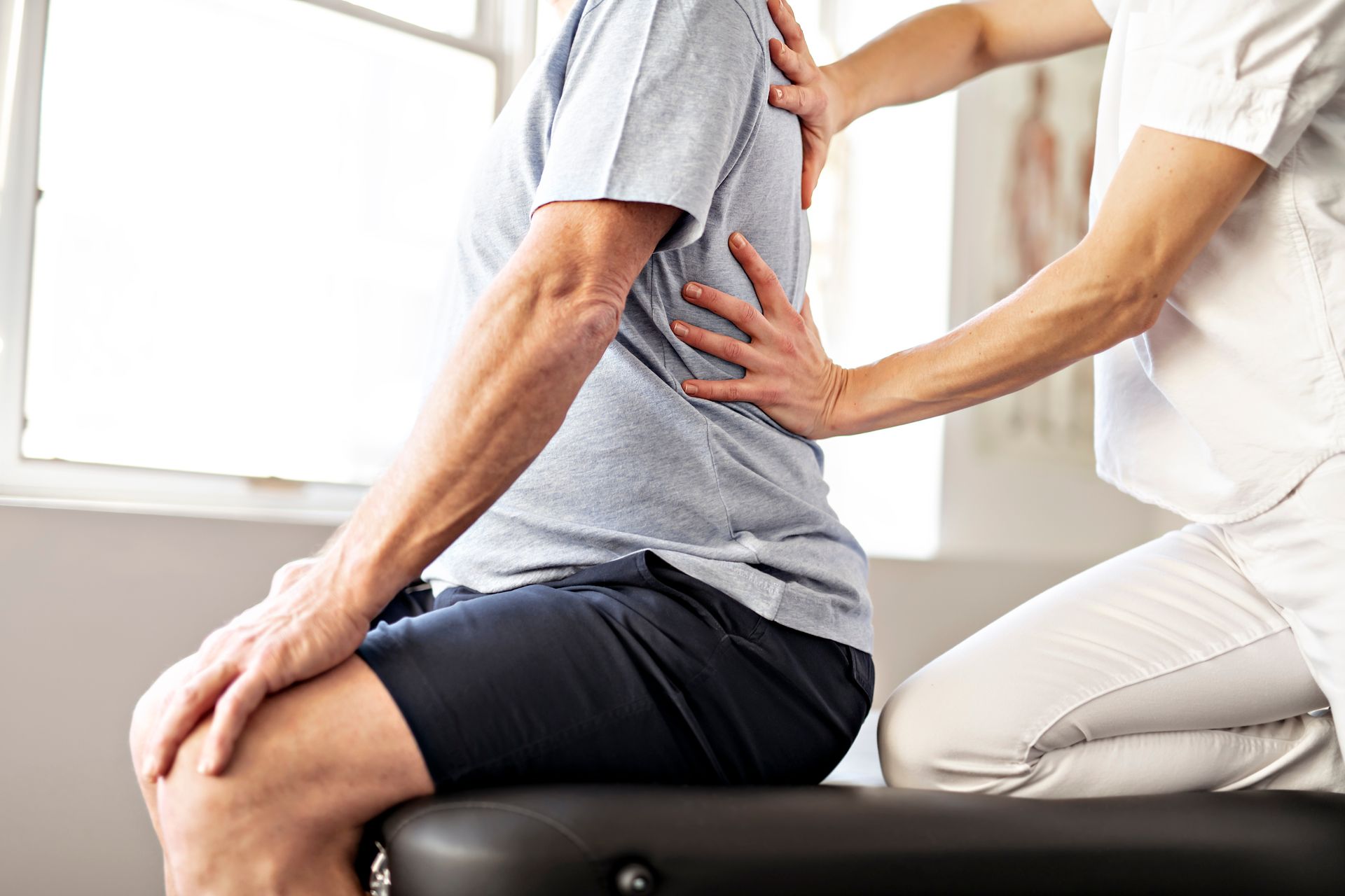 A clinician performs a physical examination of a person's lower back while they are seated on a treatment table.