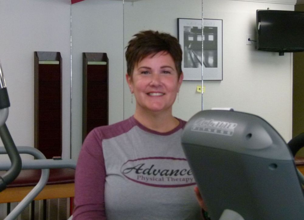 A person wearing a grey and purple shirt smiles while at a physical therapy clinic with gym equipment.
