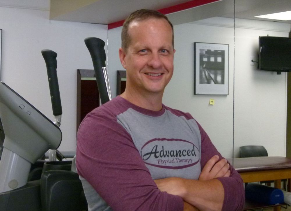 A person with a pleasant expression stands with arms crossed in a physical therapy clinic, wearing a grey and maroon shirt.