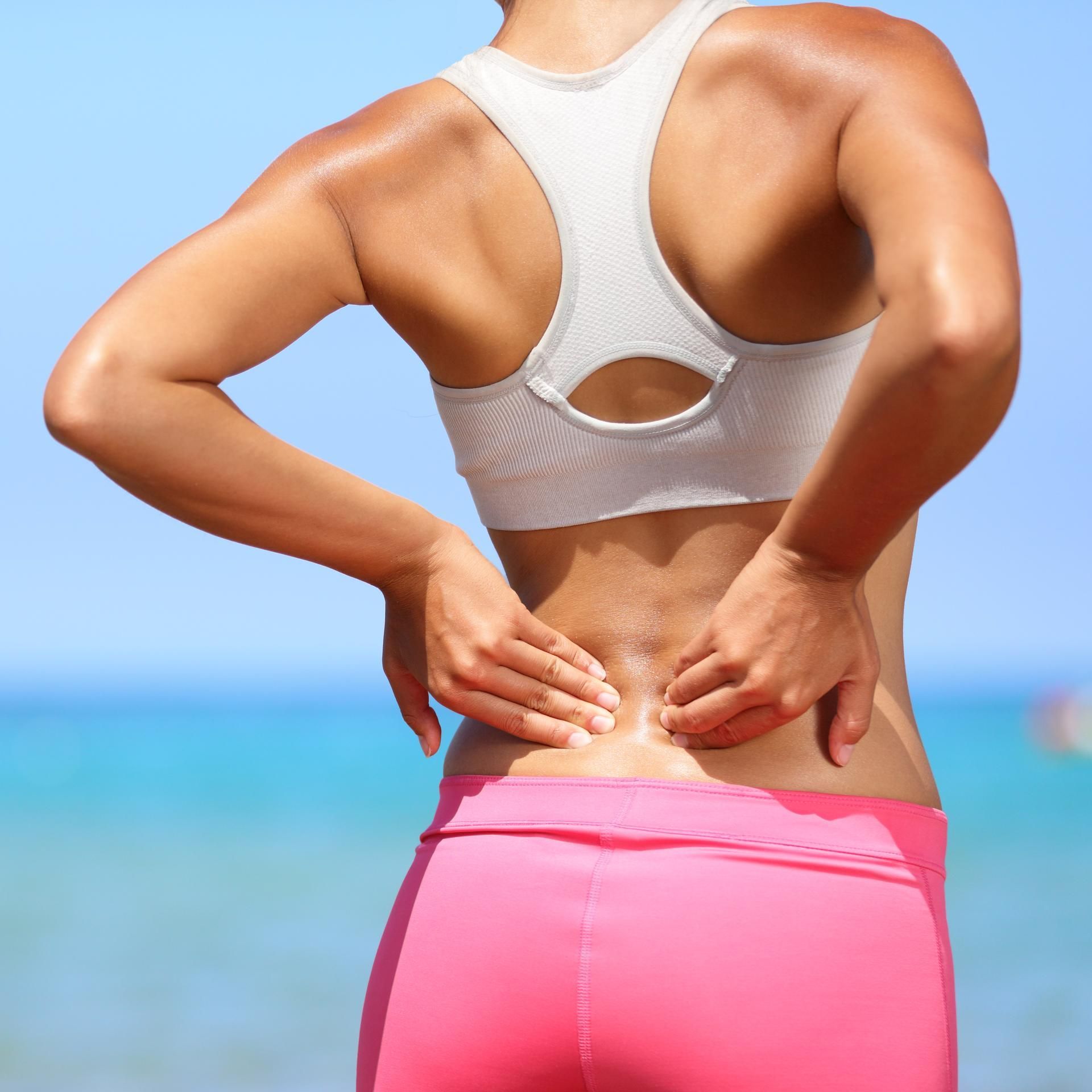 Woman in workout clothes, touching lower back. Pink pants, white sports bra. Beach background.