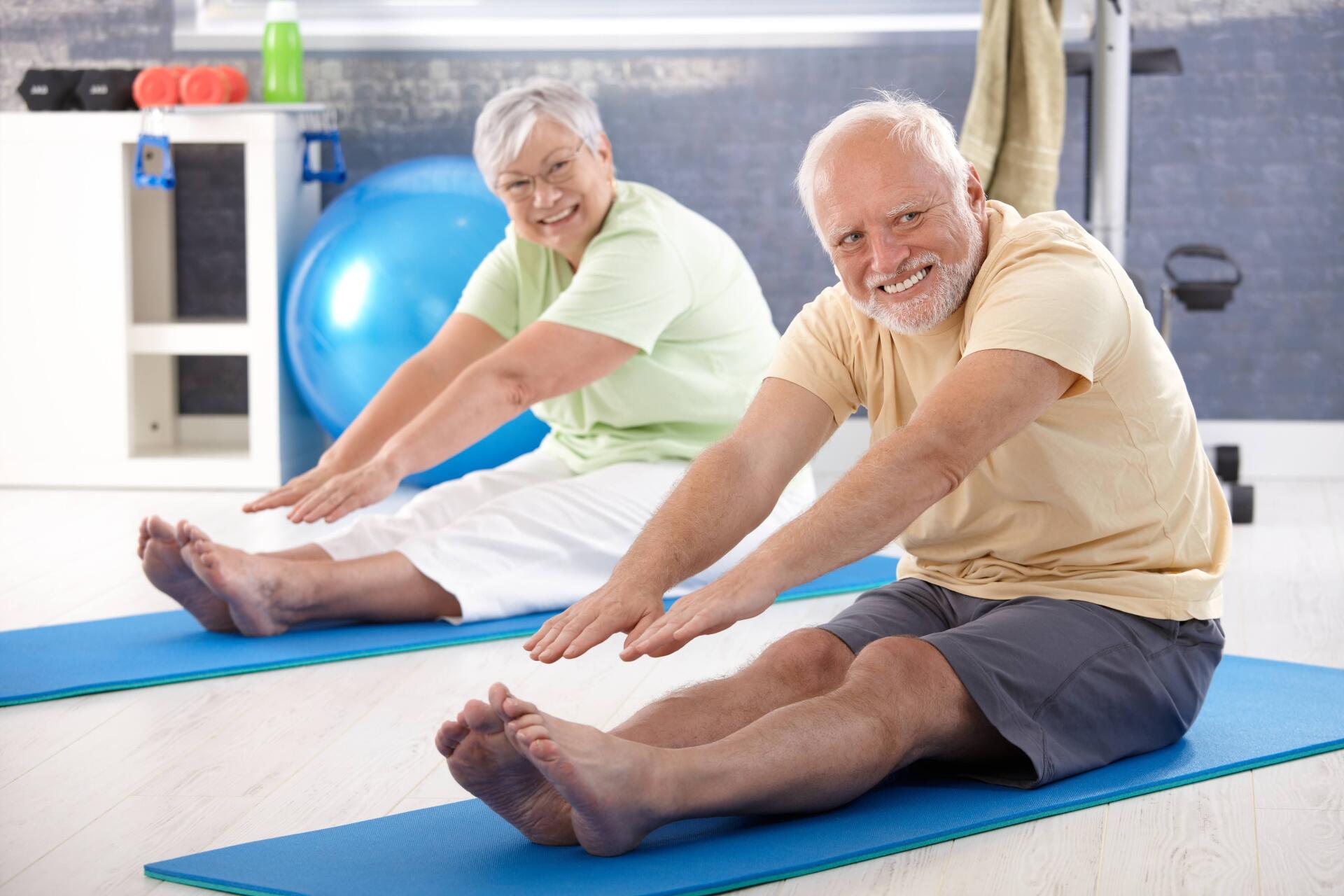Two older adults, smiling, stretching forward on blue mats in a fitness room.
