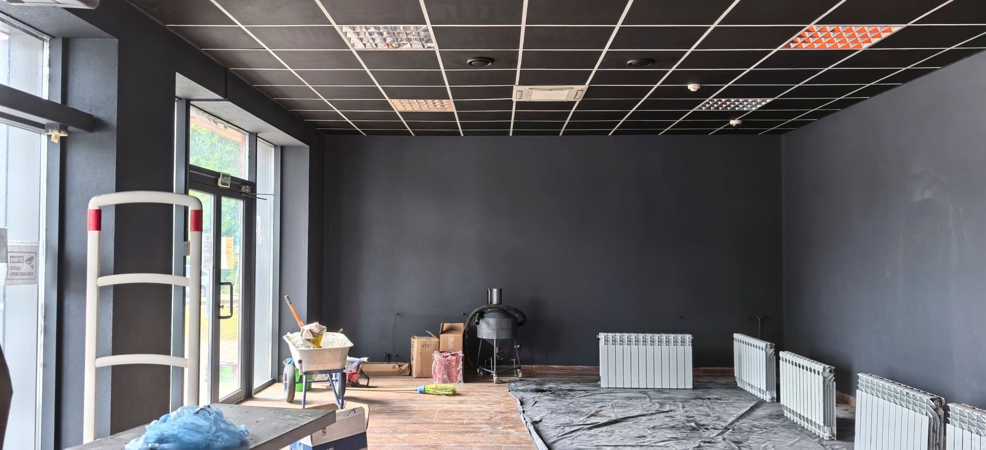 An empty, dimly lit room with dark gray walls, a black grid ceiling, large windows, construction gear, and radiators.