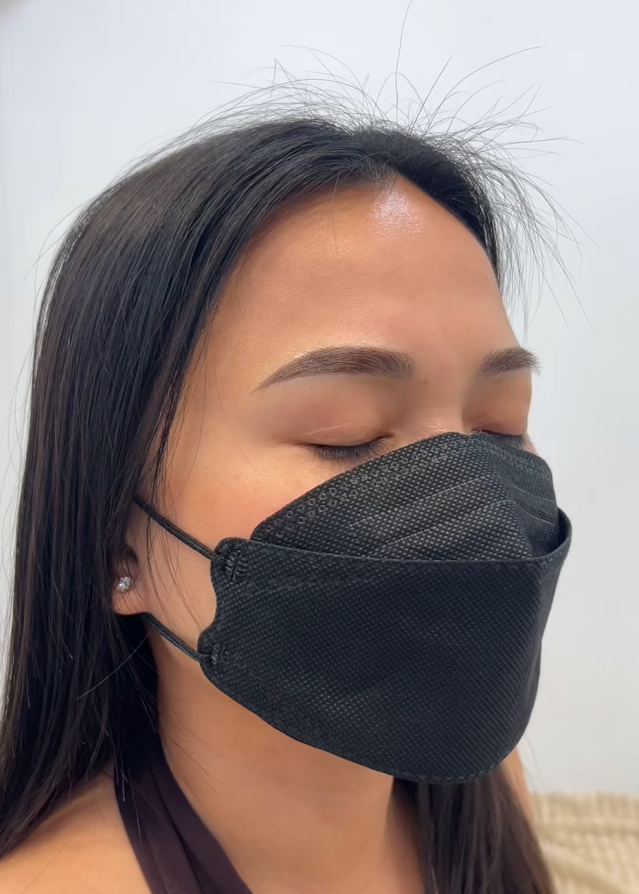 Woman with a face mask, eyeliner, and eyebrow tattoo, looking at the camera.