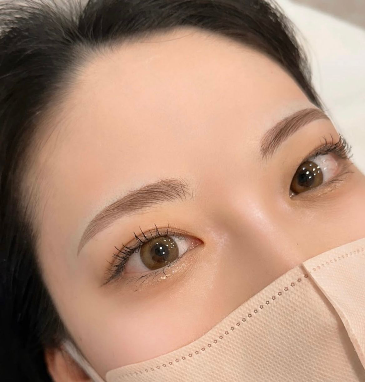 Close-up of eyes above a beige face mask, with neatly shaped eyebrows and soft makeup