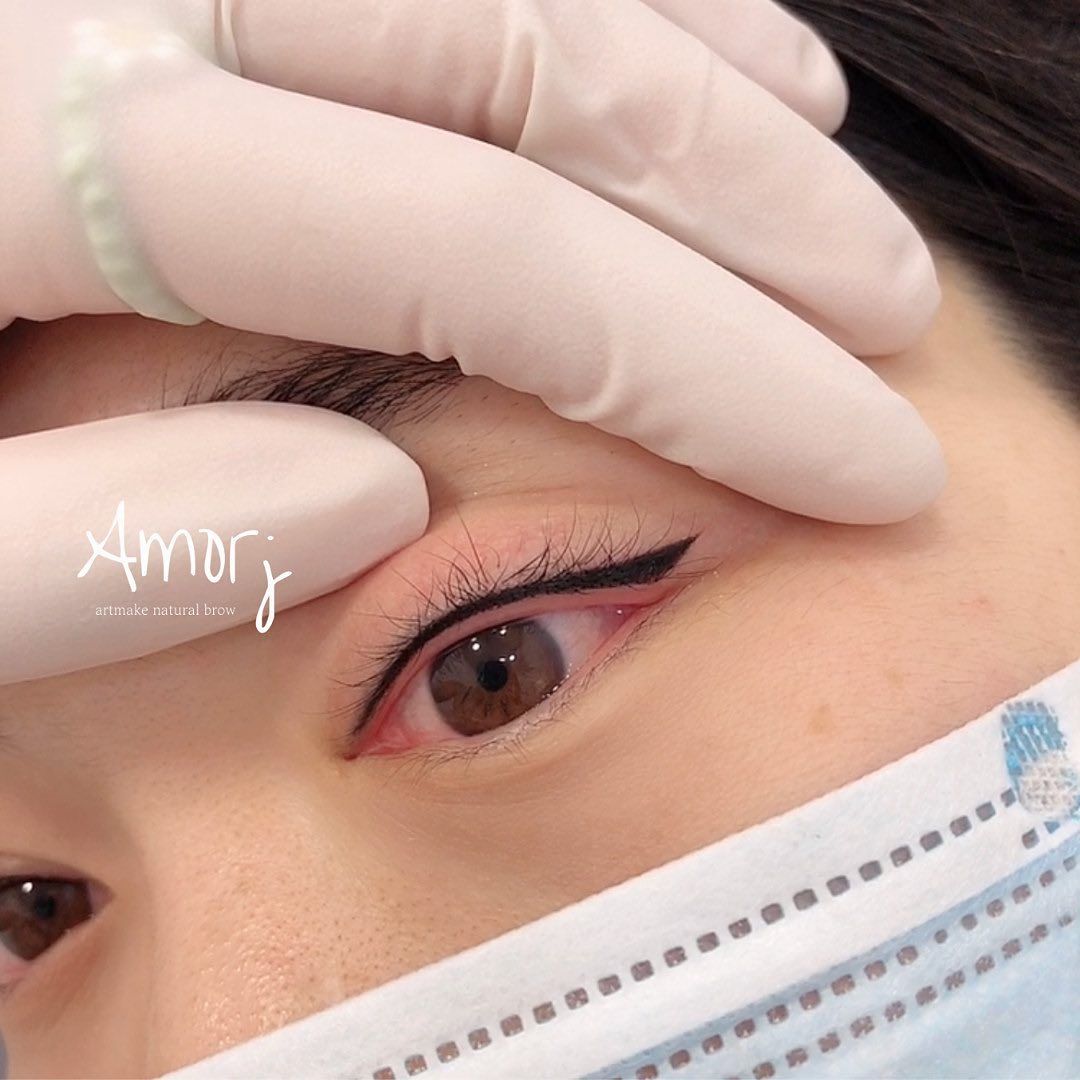 Person's eye with eyeliner tattoo. Hand holding eyelid open. Brown eye. Mask visible.