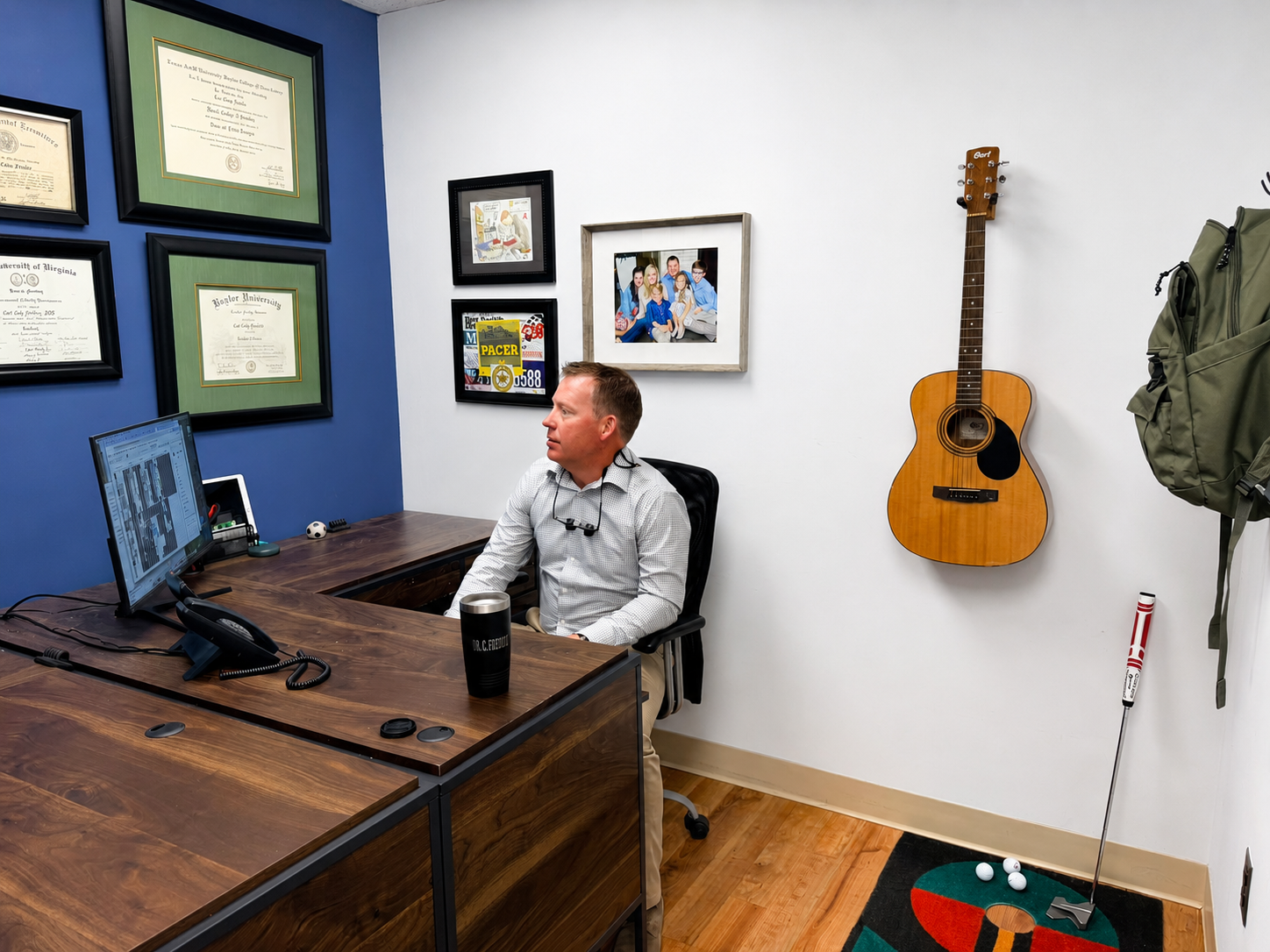 Man seated at a desk in a small office, with framed certificates on the wall and a guitar hanging nearby.