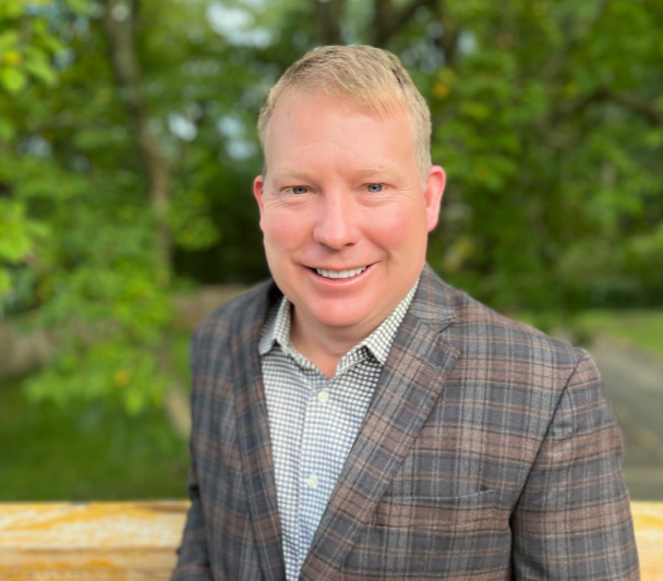 Smiling man in a plaid blazer outdoors with blurred greenery in the background.