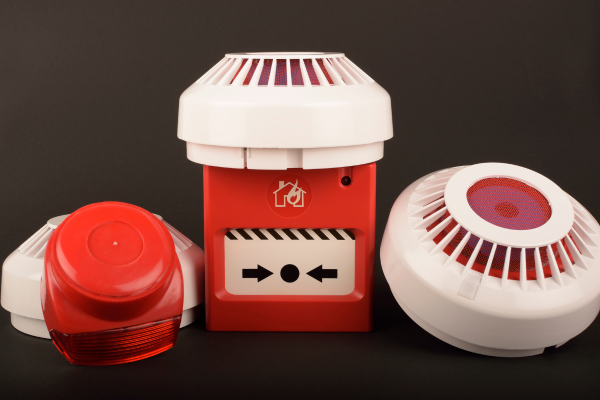 Red and white fire safety equipment: alarm, strobe light, and smoke detectors on a dark background.