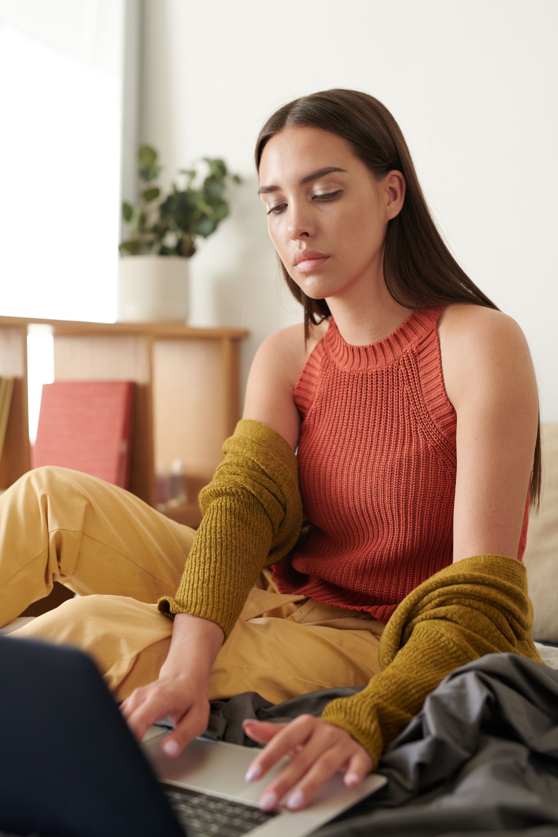 Woman in a rust sweater and mustard pants using a laptop indoors, seated on the floor with a focused expression