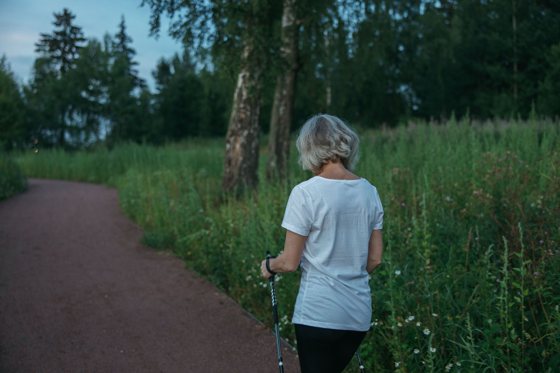 A woman walking briskly along a sunlit park path surrounded by green trees.