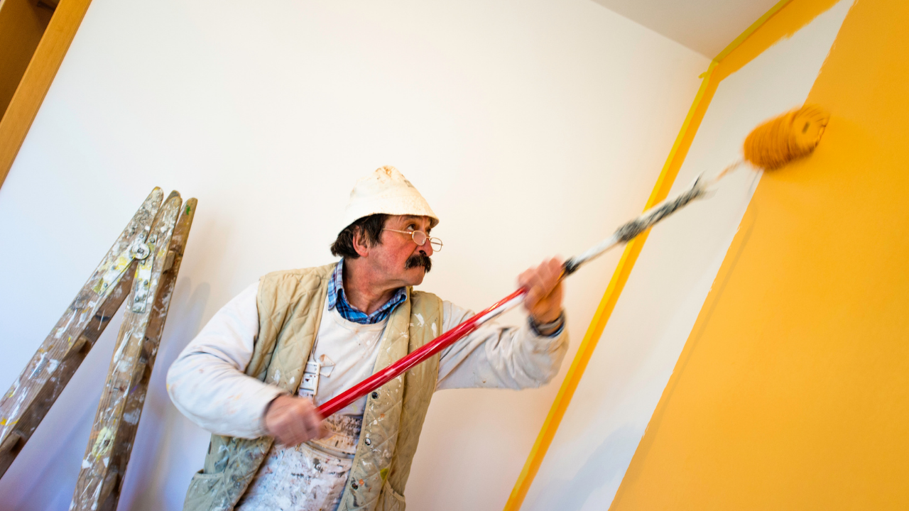 Man painting a wall with a roller. The wall is yellow and white, and he's wearing a hat, glasses, and a vest.