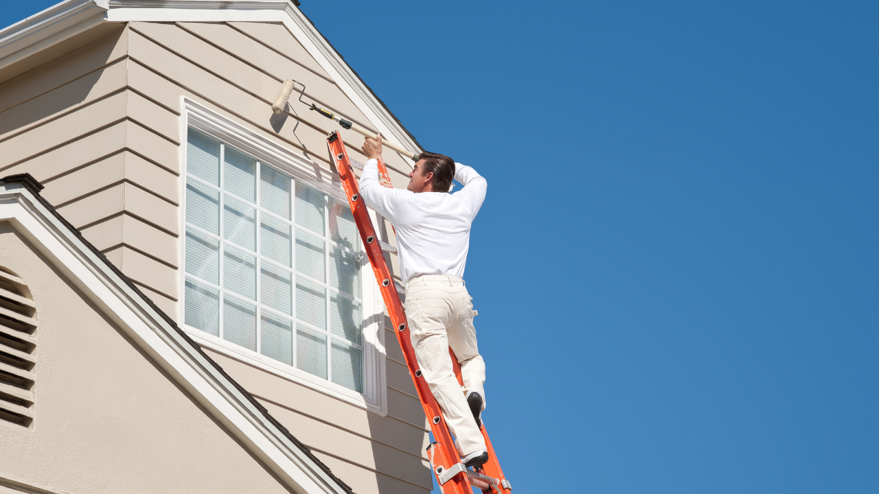 Man on a ladder painting the exterior of a house; bright blue sky.