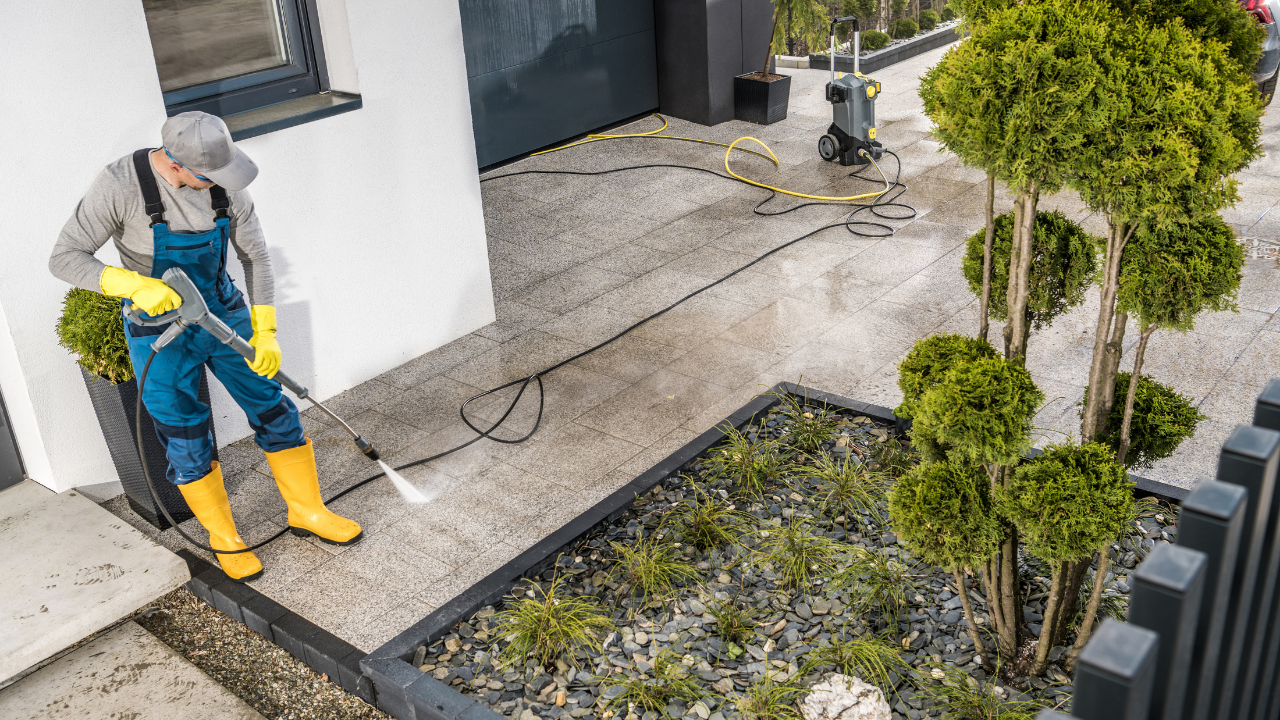 Person wearing overalls pressure washing a concrete patio next to a garden.