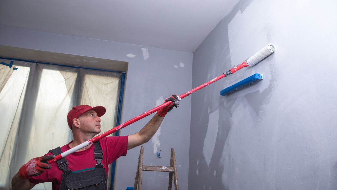 A person in red shirt and overalls paints a wall with a roller attached to a pole inside a room.