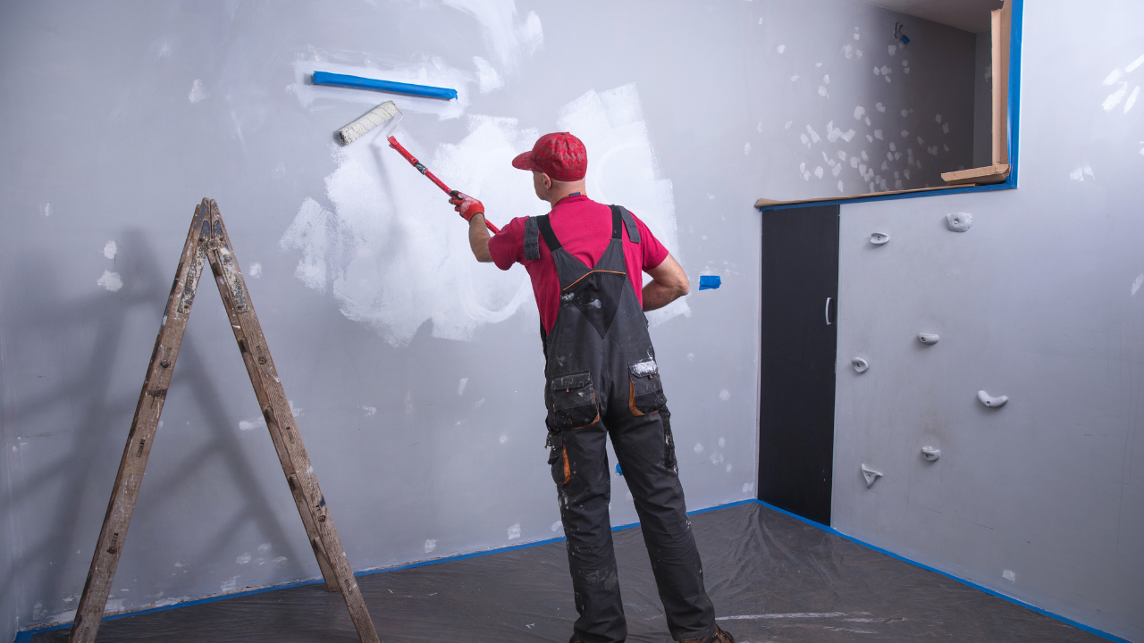 Person painting a wall with a roller. They are wearing overalls and a red hat. A ladder is nearby.