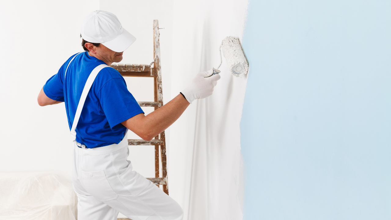 Painter in blue shirt and white overalls uses a roller to apply blue paint to a white wall near a ladder.