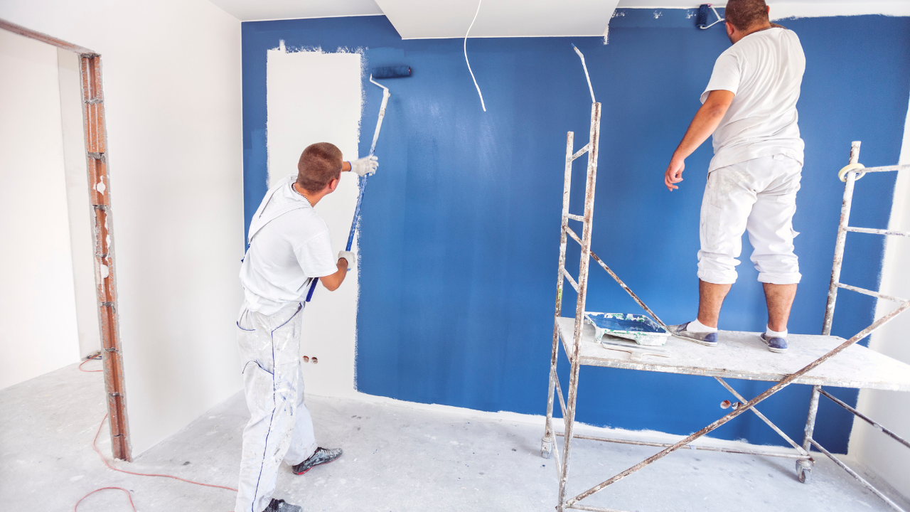 Two painters in white coveralls painting an interior wall blue with rollers; one stands on a scaffold.