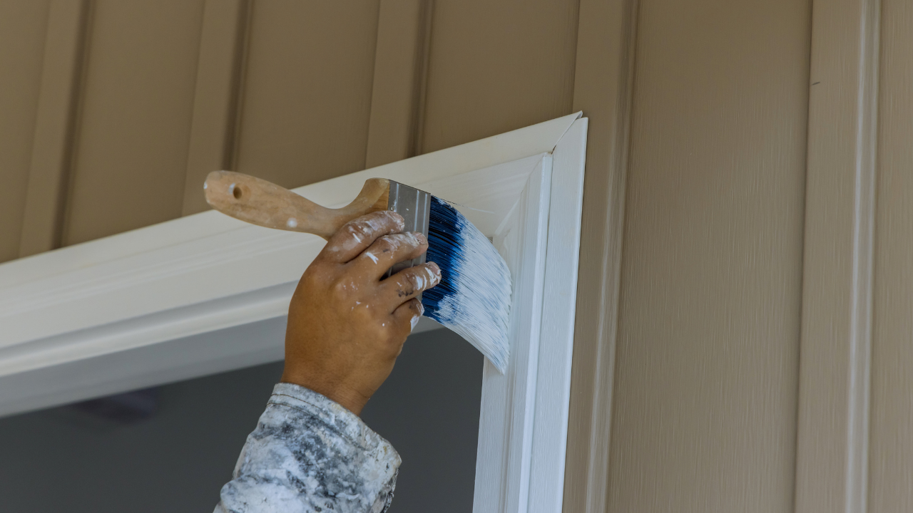 Person painting white trim around a door with a brush, against tan siding.