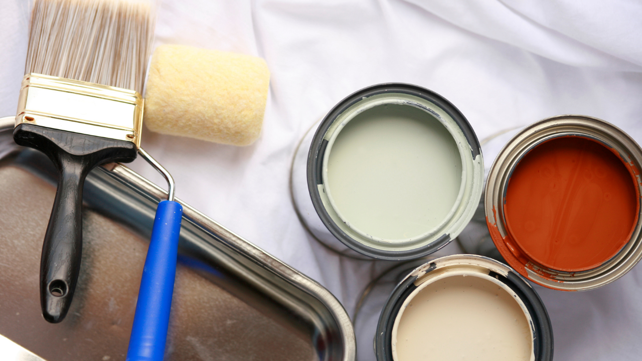 Paint supplies on white fabric: brushes, roller, paint tray, and open cans in green, orange, and cream colors.
