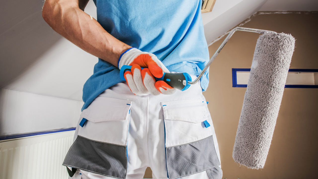 Painter in white and blue work clothes holding a paint roller behind their back, indoors.