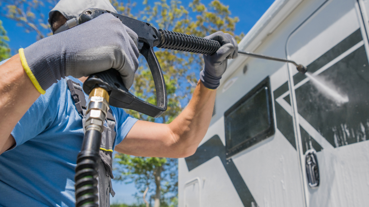 Person wearing gloves pressure washing RV exterior.