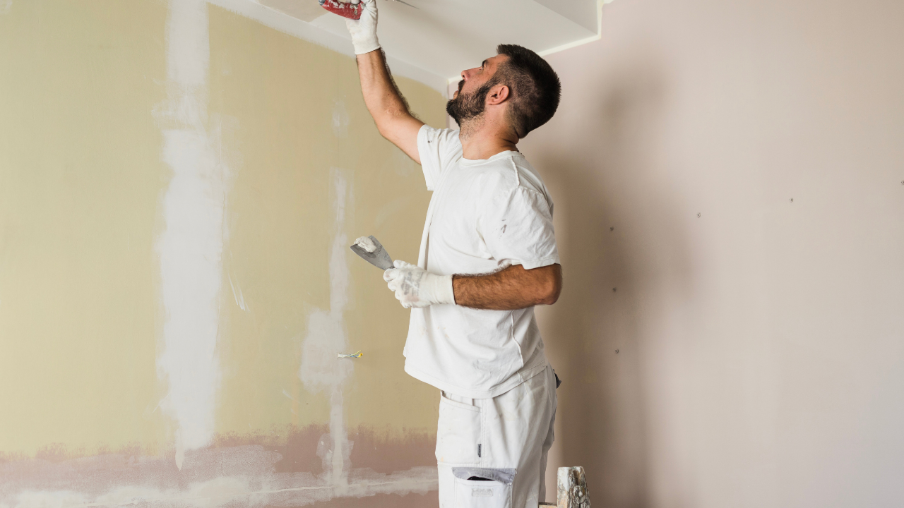 Man in white overalls, gloves, sanding a ceiling corner with tools, light-colored walls.