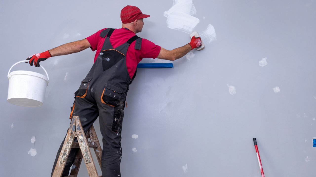 Painter on a ladder applies plaster to a gray wall, holding a paint bucket and roller.