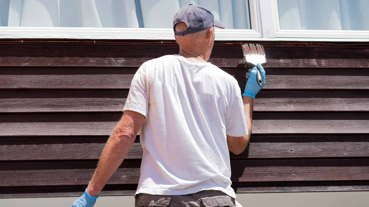 Person painting a brown exterior wall white near a window, wearing a cap and gloves.