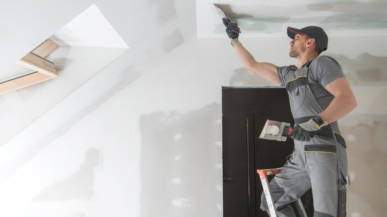 Man on a ladder applying drywall compound to a ceiling with a trowel; interior setting.